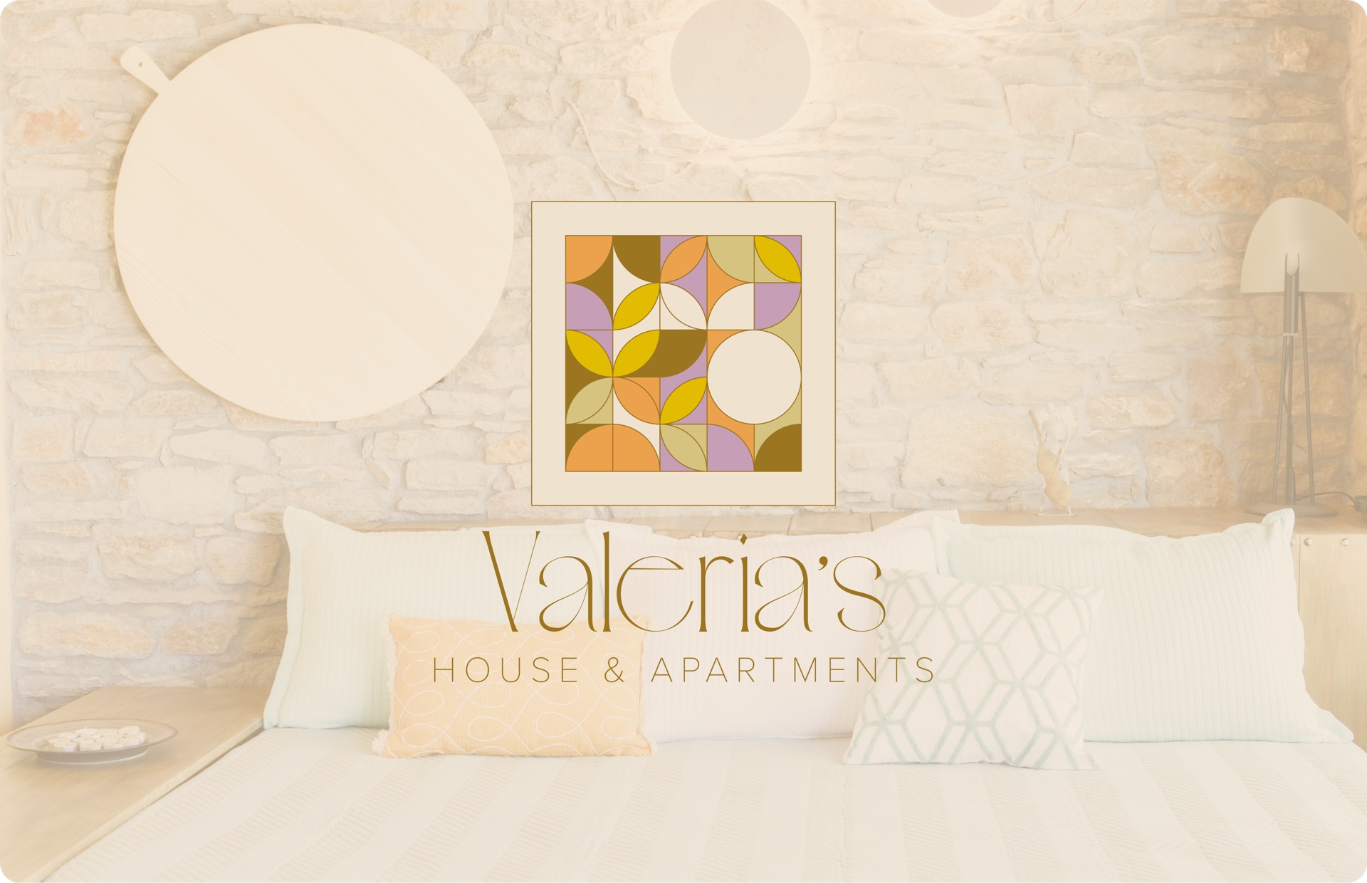 Valeria's House mosaic logo on a Cycladic stone bedroom wall above a softly dressed double bed with linen pillows