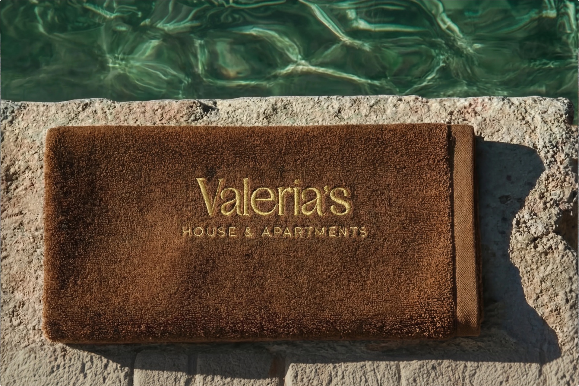 Embroidered brown bath towel reading 'Valeria's House — House & Apartments' laid on the limestone pool deck above clear emerald water