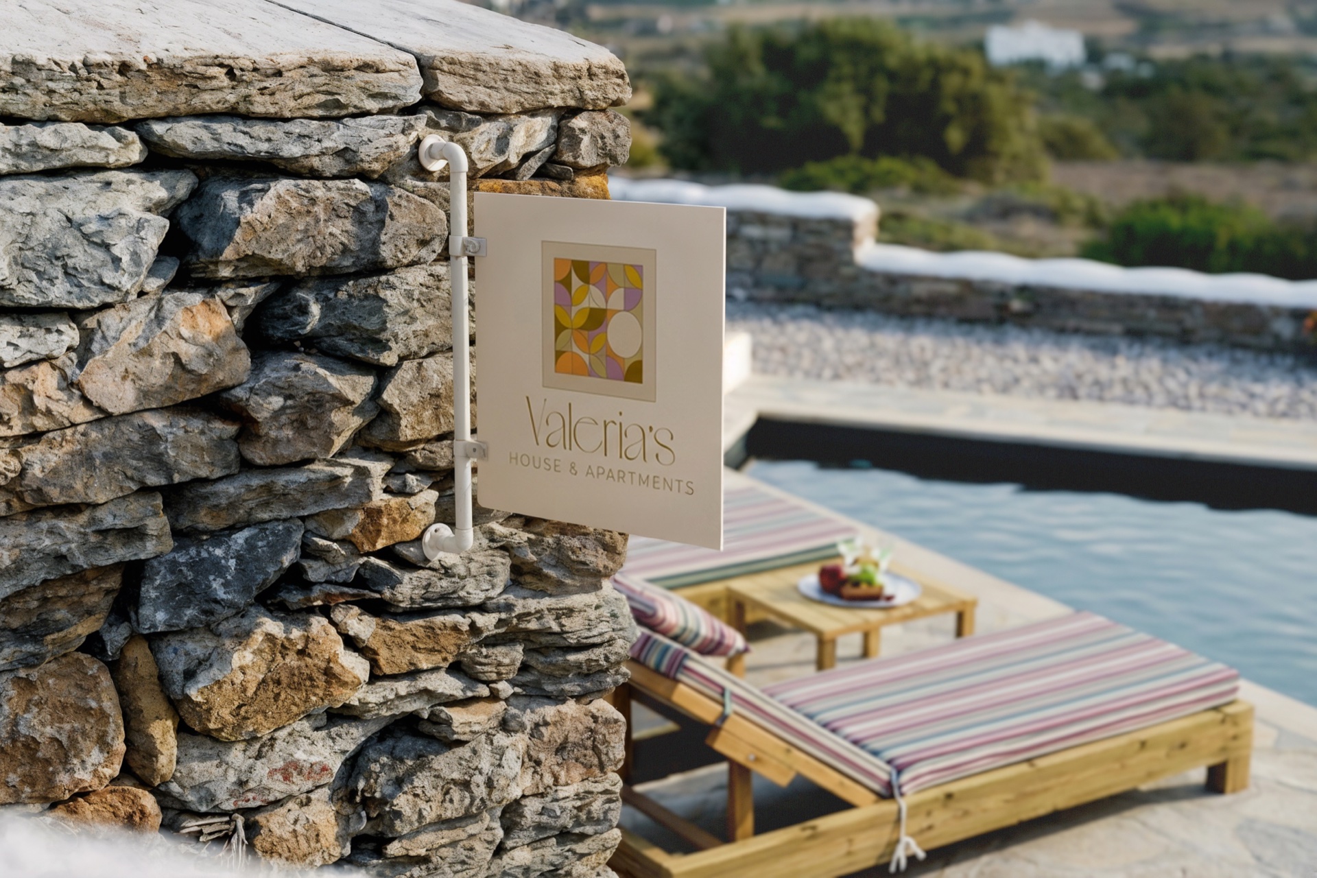 Valeria's House mosaic plaque mounted on a Cycladic stone garden wall, with striped loungers and the open-air pool deck behind